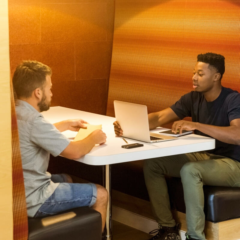 Two people in casual clothing sitting opposite in a booth. They look like they are in a work meeting - one has a laptop and the other has a sheet of paper & pen