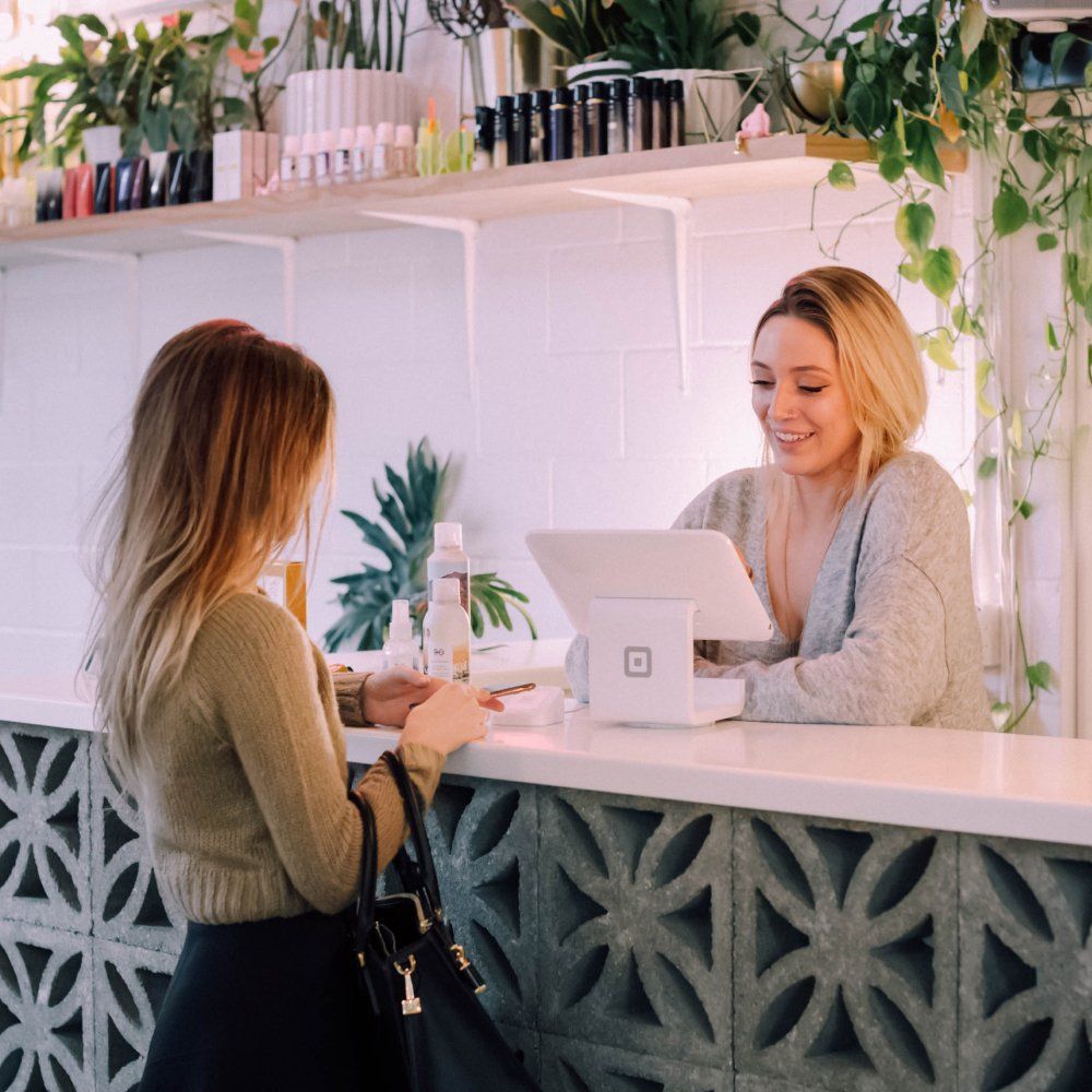 Femme debout devant une étagère de produits de beauté et servant une cliente à la caisse.